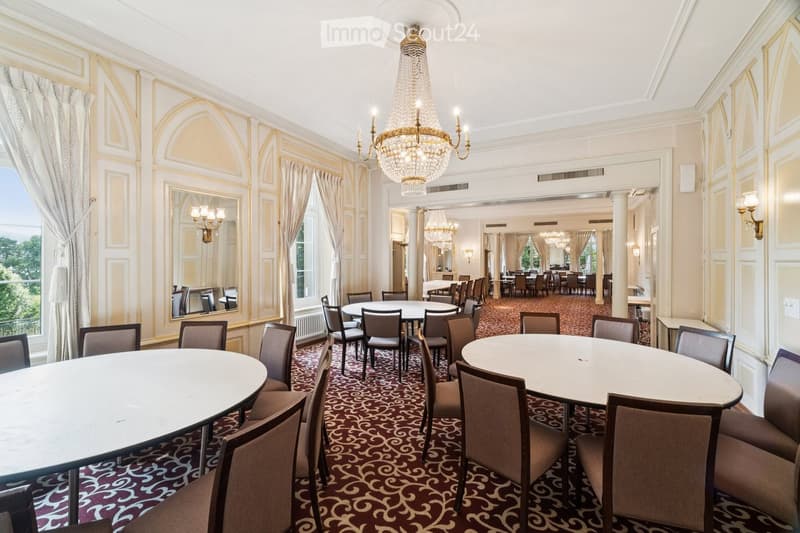 Elegant dining area with round tables, chairs, ornate chandelier, large windows with curtains, and a patterned carpet.
