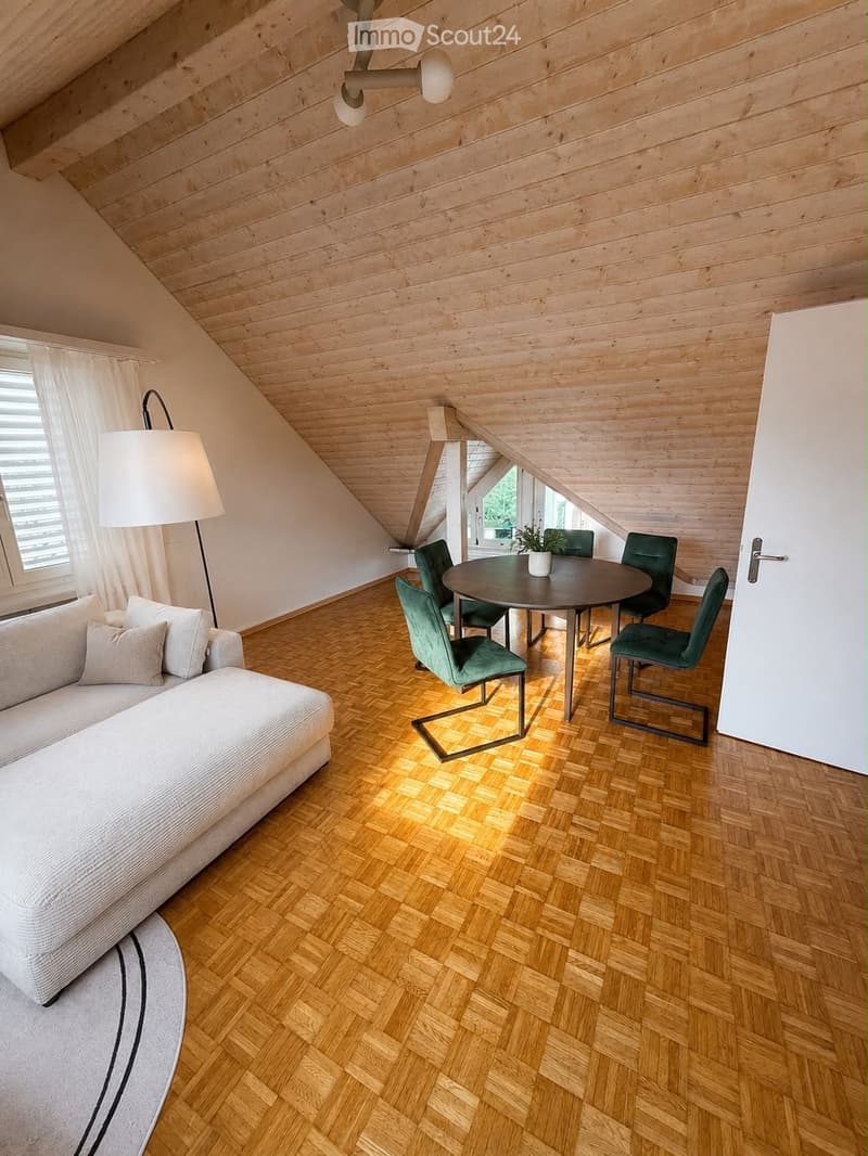 Modern attic room with wooden slanted ceilings, herringbone wooden flooring, a white sofa, a round dining table with green chairs, a floor lamp, and a potted plant on the table.
