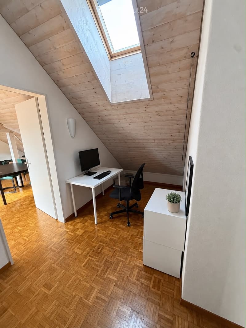 A modern, well-lit attic room with wooden herringbone flooring, white walls, and a slanted wooden ceiling. Features include a desk with a computer, a comfortable office chair, a small side table with a potted plant, and a large skylight providing natural