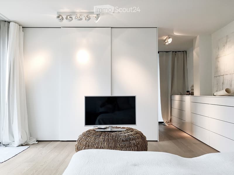 Modern bedroom with a large bed, wicker stool, flat-screen TV, built-in wardrobe, and a small en-suite bathroom with a mirror and drawer unit.