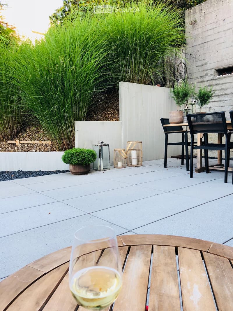 A modern outdoor patio with a wooden table, black chairs, a glass of white wine, and decorative plants including tall grasses and small shrubs.