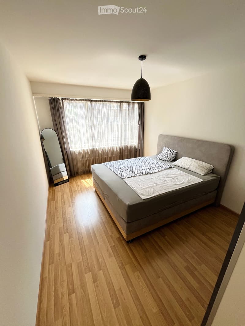 A modern bedroom with a wooden floor, a grey upholstered bed, a pendant light, and a large window with curtains.
