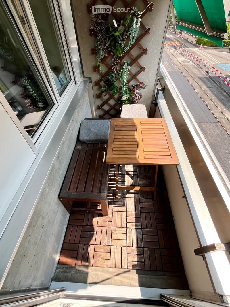 Balcony with wooden decking, a small table, two chairs, and a stool, with a decorative wooden trellis and green plants.