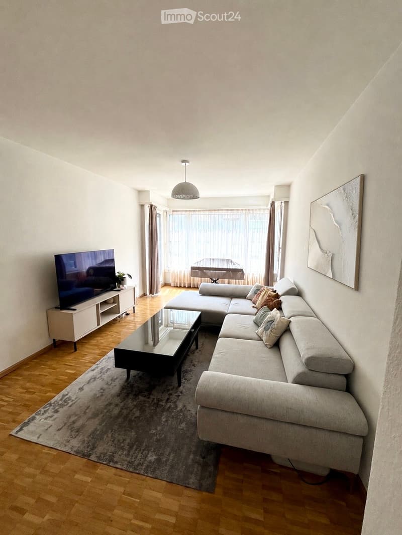 Modern living room with light-colored walls, wooden flooring, and large windows. Features a grey sectional sofa, glass coffee table, flat-screen TV on a white stand, and a small side table with a plant.
