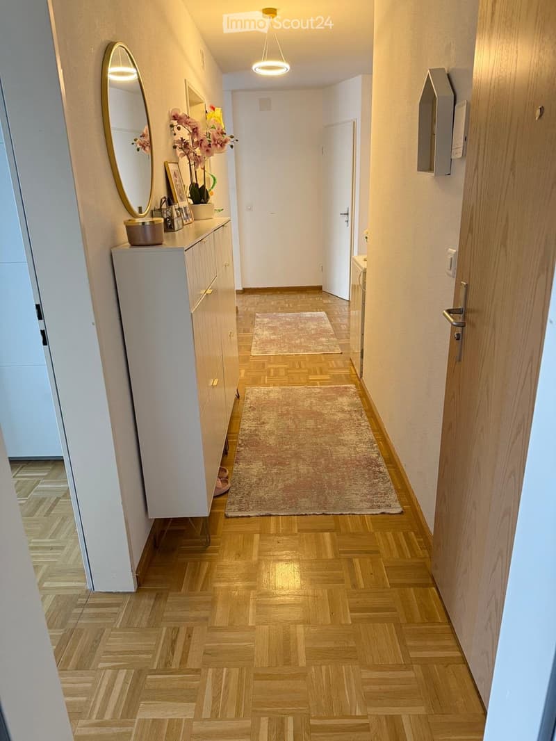 A well-lit hallway with wooden flooring, two small rugs, a small cabinet with a mirror, and decorative items on top.