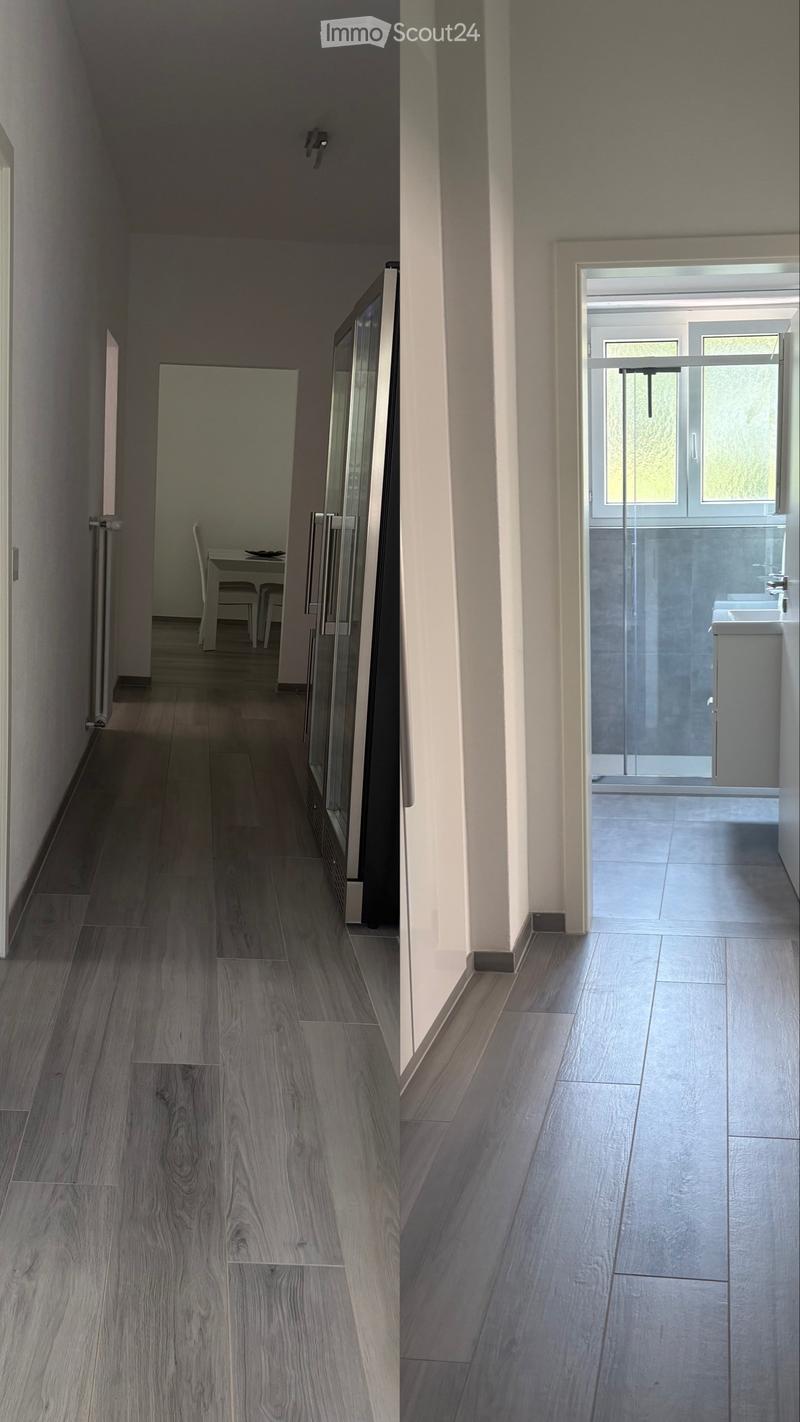 Modern hallway with wooden flooring, glass doors, and a small dining area visible at the end. The hallway leads to a bathroom with a glass-enclosed shower and a sink.