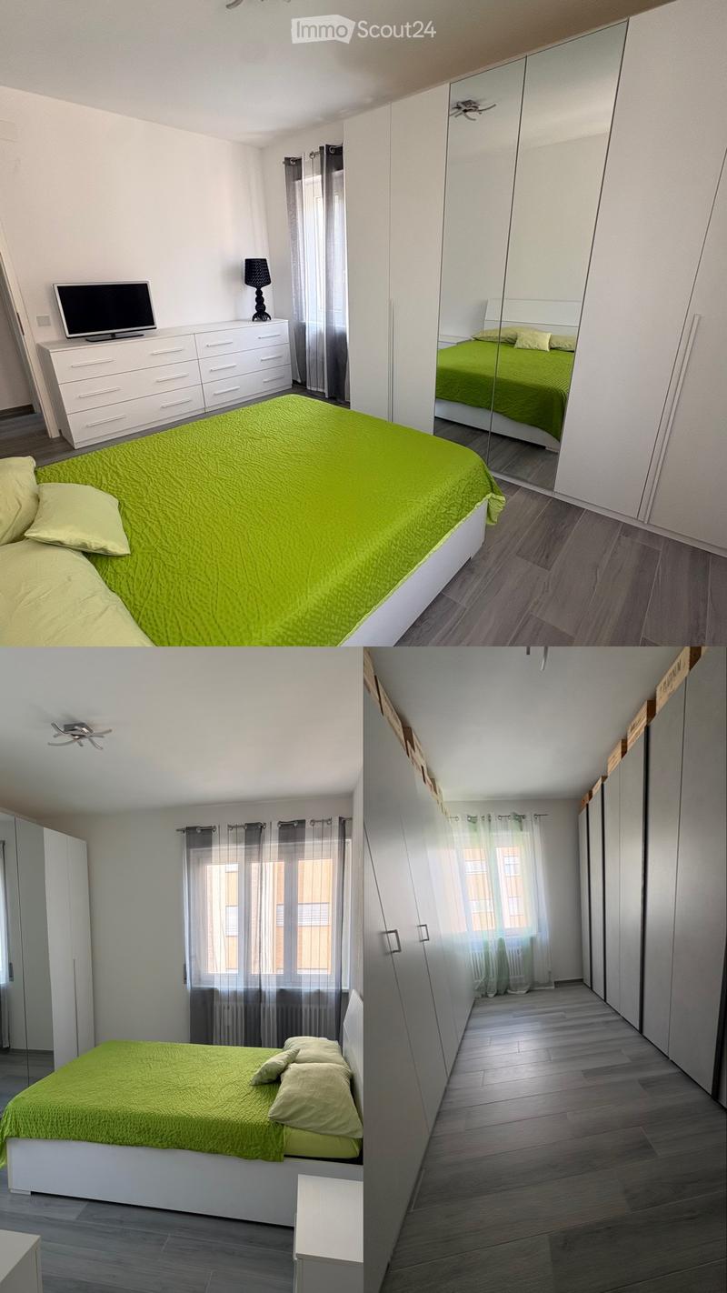 Modern bedroom with a green duvet, white nightstand, lamp, wardrobe with mirrored doors, and television on a dresser.