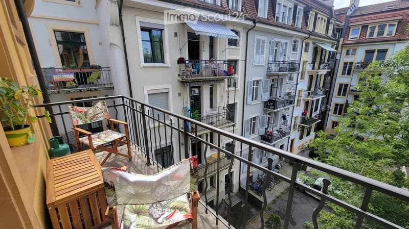 Balcony with wooden table, two floral-patterned chairs, and a potted plant, overlooking a cityscape with multiple buildings and balconies.
