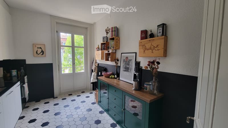 Modern kitchen with teal cabinets, wooden countertop, and black and white hexagonal tiles. Features a framed picture, vase with dried flowers, coffee maker, and shelves with decorative items.