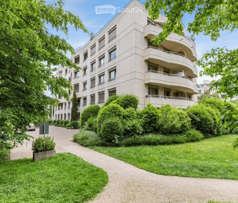 Modern multi-story apartment building with balconies, surrounded by well-maintained greenery and a paved pathway.