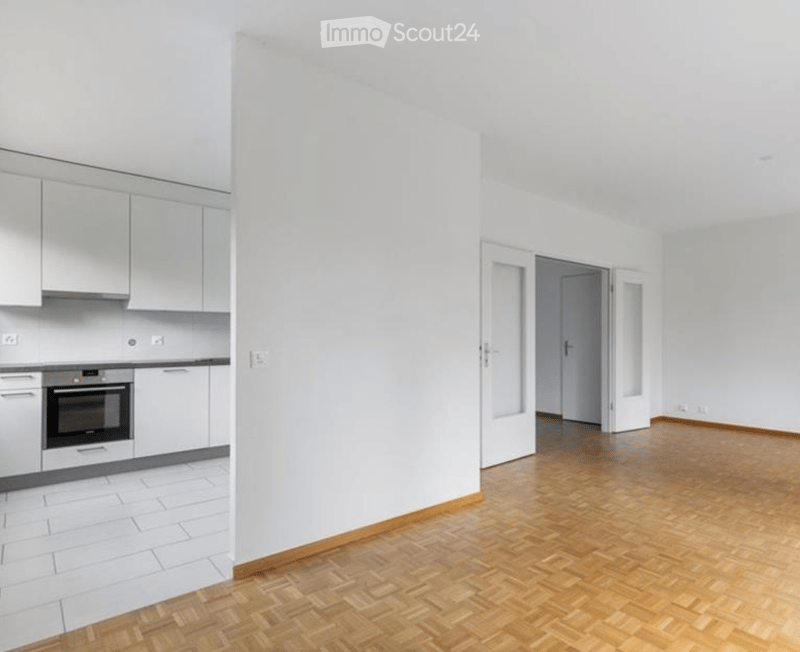 Empty room with white walls, wooden floor, and a kitchen area with white cabinets and appliances visible in the background.