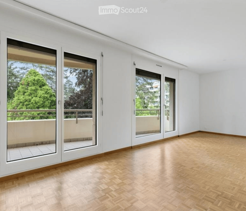 Empty room with wooden flooring, white walls, large sliding glass doors leading to a balcony with a view of greenery.