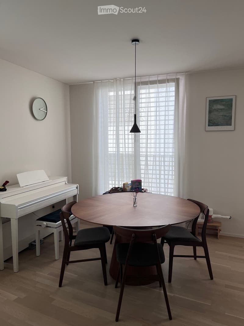 A modern dining area with a round wooden table surrounded by four chairs, a white piano, and a clock on the wall.