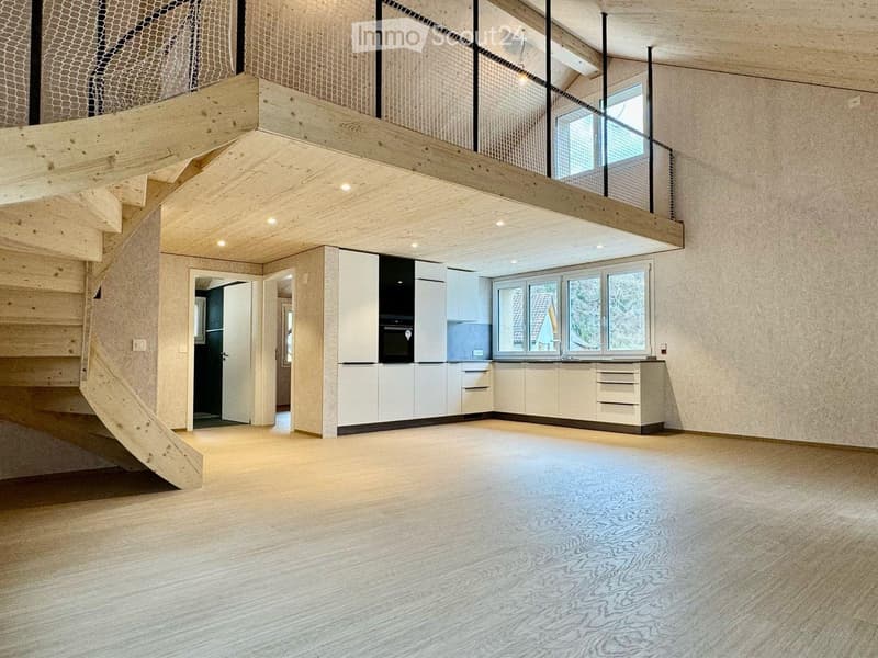 Open-plan living space with a modern kitchen area featuring white cabinets, an oven, and a sink, connected to a wooden staircase leading to an upper level with a balcony.
