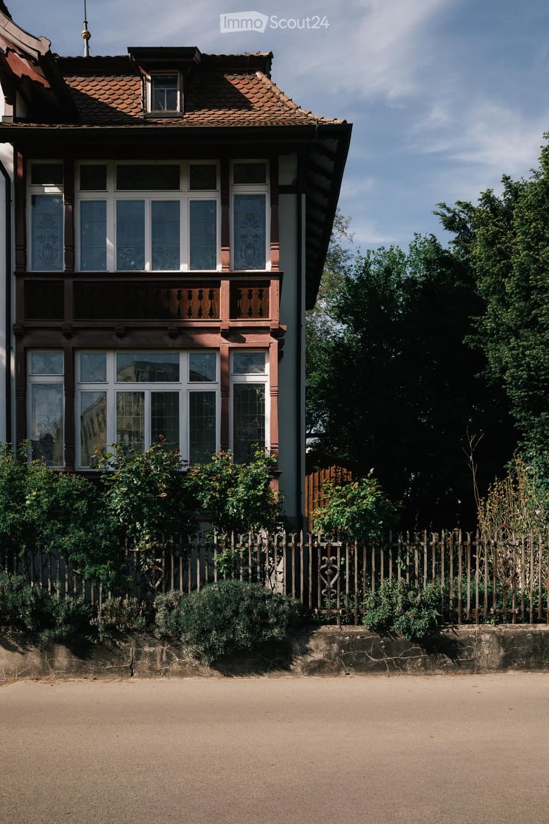 Two-story house with red-tiled roof, white-framed windows, decorative wooden balcony, and surrounding greenery.