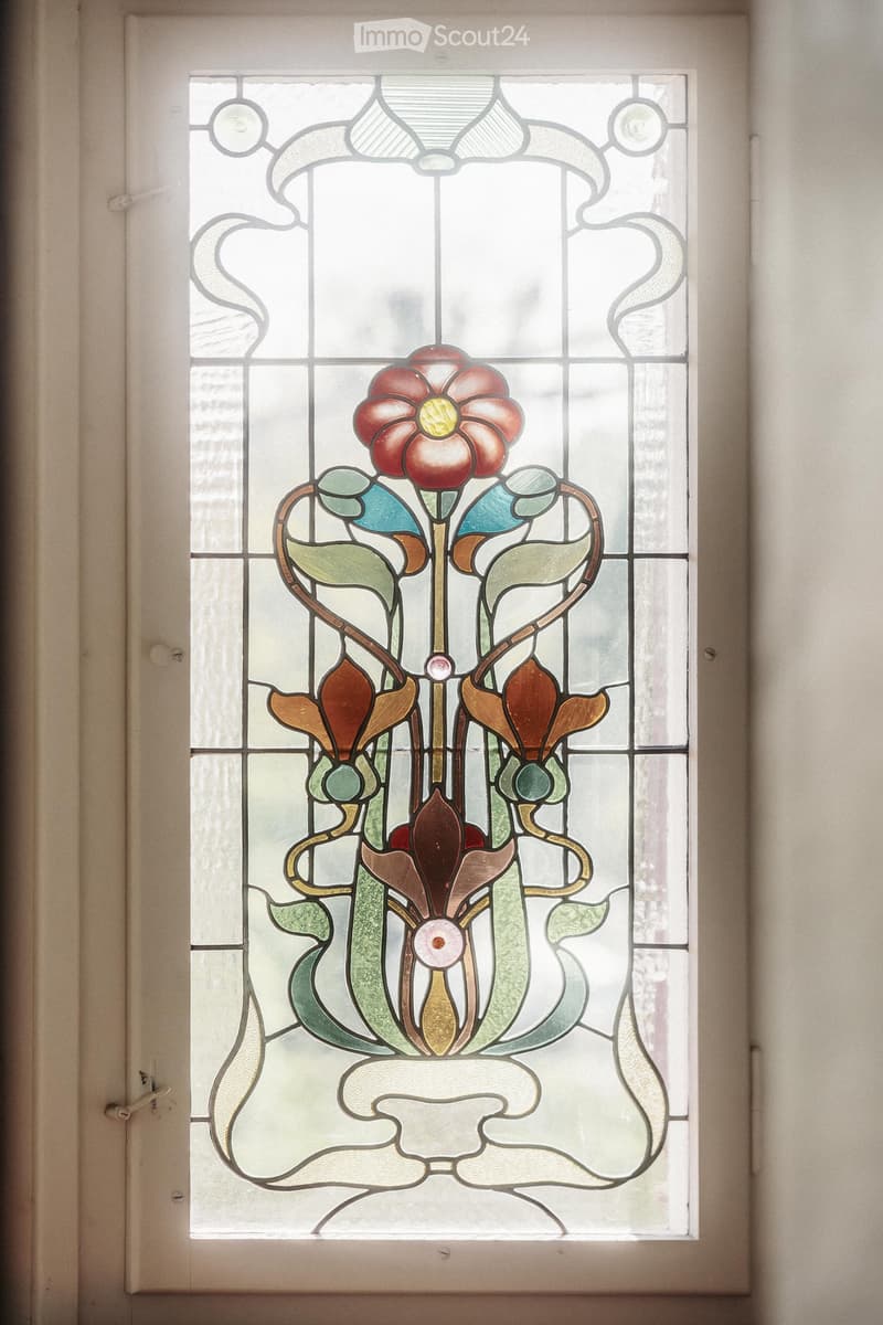 A stained glass window featuring a floral design with a central red flower and green leaves, set within a white frame.