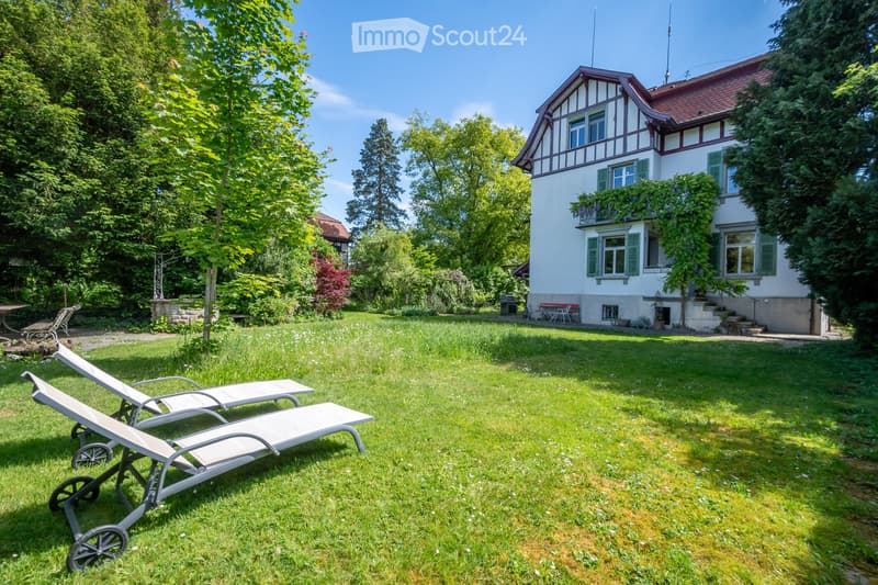 A well-maintained garden with a two-story house in the background, featuring sun loungers, a barbecue, and lush greenery.