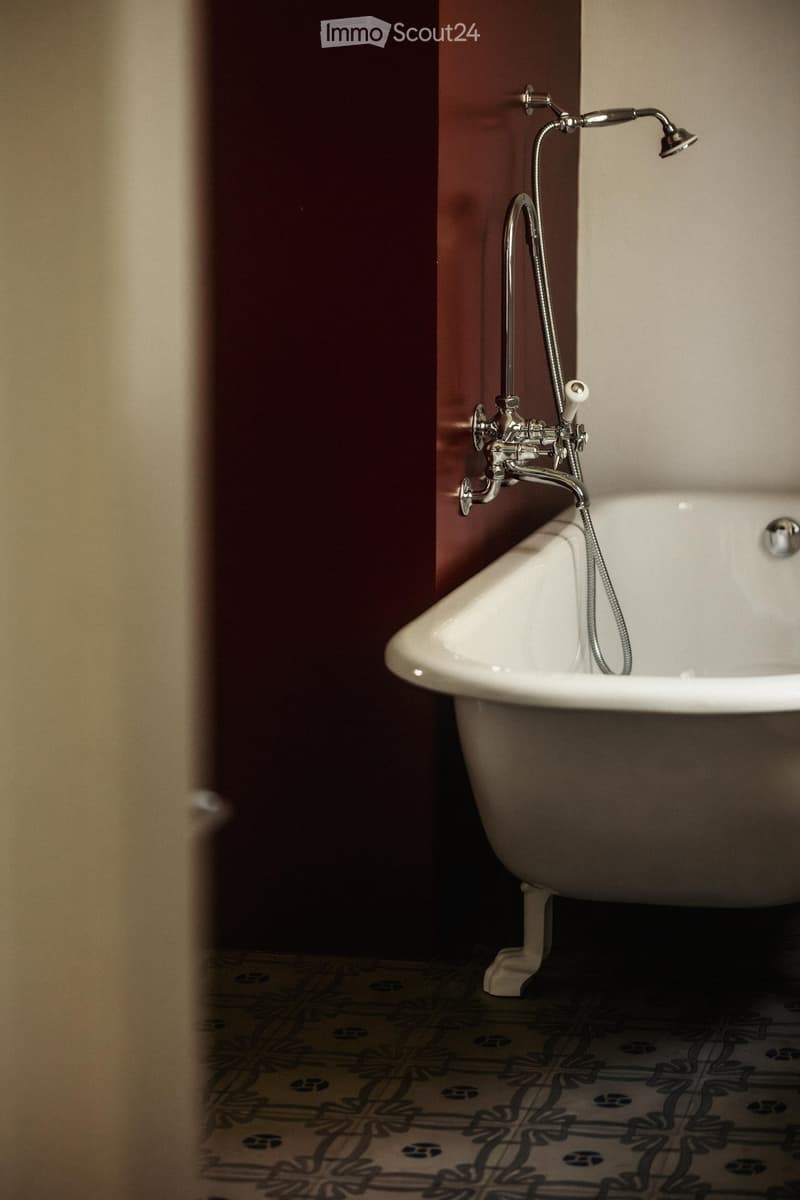 Bathtub with a modern faucet and handheld showerhead, placed against a dark red wall with patterned tile flooring.