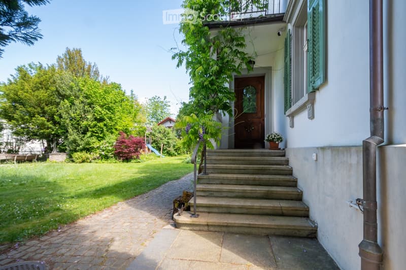 A two-story house with a raised ground floor, featuring a stone staircase leading to a wooden front door, surrounded by a well-maintained garden with trees and shrubs.