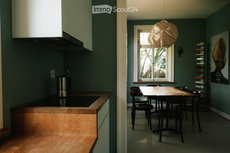 A modern kitchen and dining area with dark green walls, a wooden countertop, a pendant light, and a dining table with chairs.
