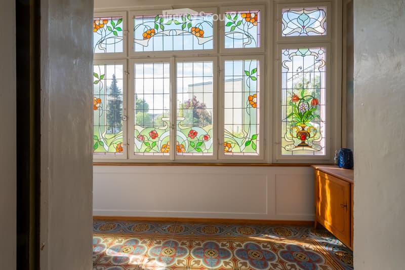 An empty room with colorful stained-glass windows, a decorative tile floor, and a small wooden cabinet with a vase on top.