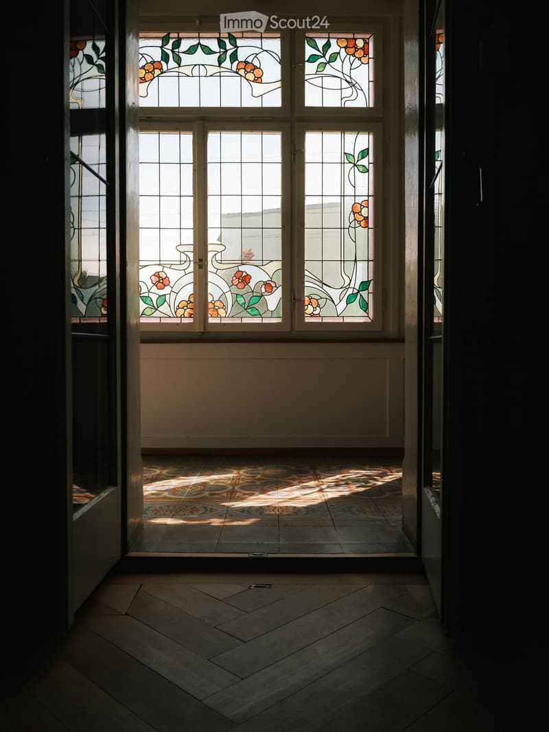 Hallway with stained glass windows, patterned tile floor, and wooden flooring leading to another area.