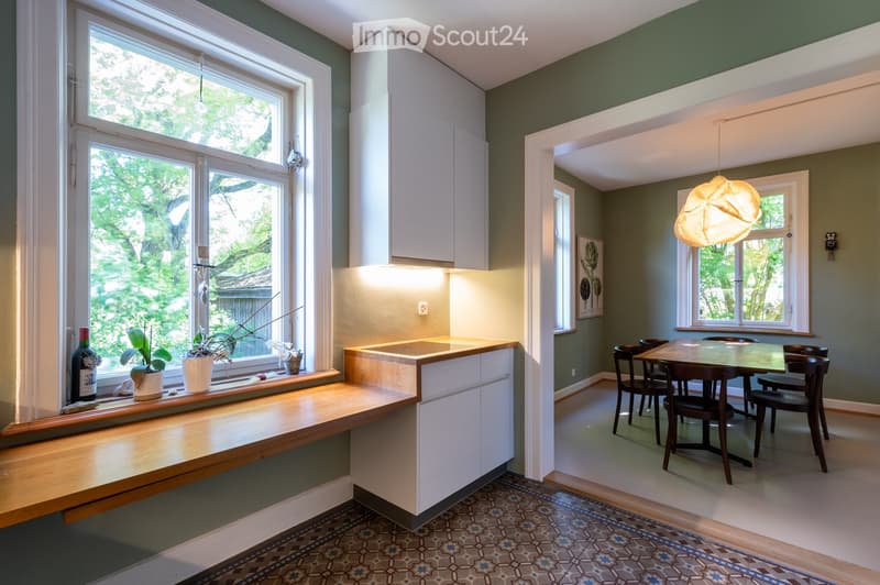 Bright kitchen with white cabinets, a wooden countertop, and a dining area with a table and chairs. The room features large windows with a view of greenery outside, and decorative items on the windowsill.