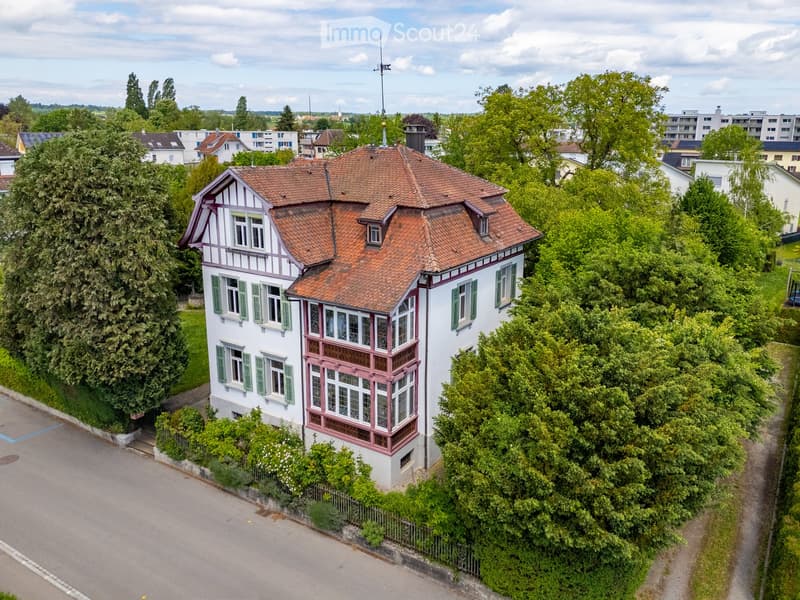 A two-story house with a red-tiled roof, white walls, green shutters, and decorative pink trim around the windows and roofline. The house is surrounded by lush greenery and trees, with a paved driveway and neighboring houses visible in the background.