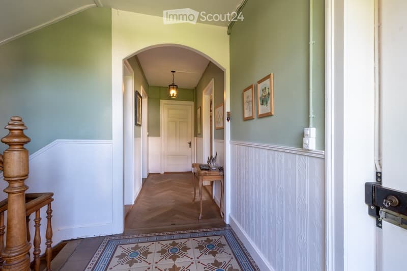 Elegant hallway with wooden herringbone flooring, decorative patterned tiles, framed artwork, and a small wooden table with items on it.