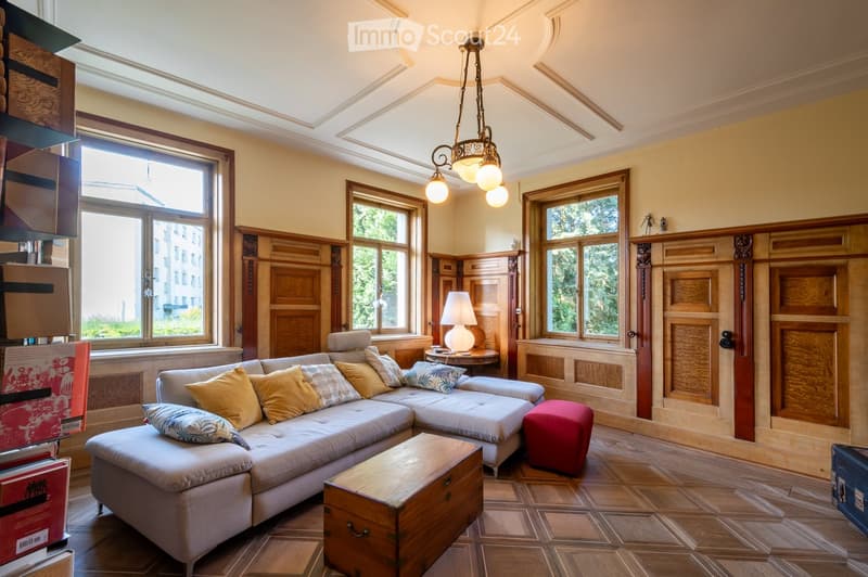Elegant living room with a large sectional sofa, colorful cushions, a wooden coffee table, a red ottoman, and a table lamp on a side table. The room features wooden paneling, large windows, and a decorative chandelier.