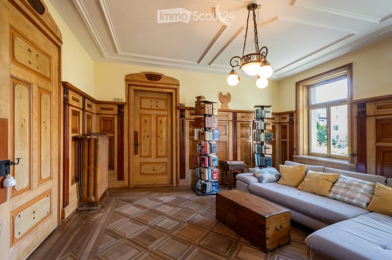 Elegant living room with wooden floors, large windows, a grey sectional sofa, wooden coffee table, bookshelves, and ornate wooden cabinetry.
