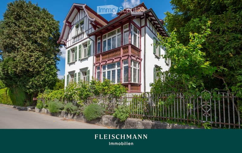 A two-story house with white walls, red trim, and green shutters. The house features a prominent balcony with ornate railings and large windows. The property is surrounded by lush greenery and a wooden fence.