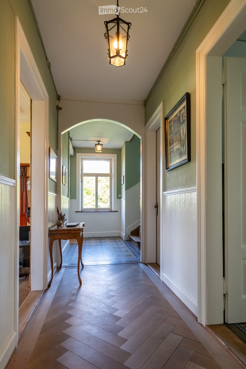 Elegant hallway with herringbone wooden flooring, arched doorway leading to another room, framed artwork on the wall, small wooden table with decorative items, and a hanging lantern-style light fixture.