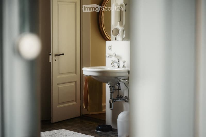 A small bathroom with a white sink, chrome faucet, round mirror, and a closed door visible in the background. A towel hangs on the wall, and a small trash can is placed on the floor near the sink.