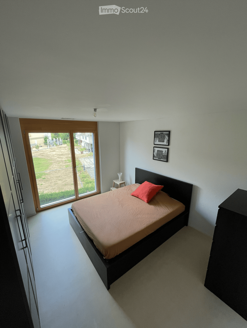 Modern bedroom with a bed, nightstand, and picture frames on the wall. Large window with a view of the outdoor area.
