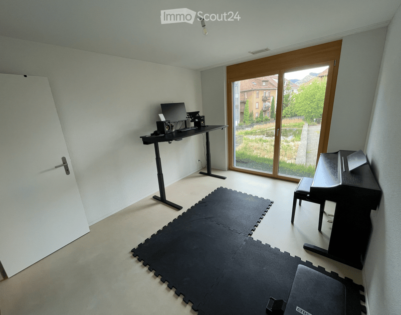 A modern, minimalist room with a standing desk, laptop, keyboard, mouse, speakers, and a stool. The room features a large sliding glass door with a view of greenery and buildings outside, and a black exercise mat on the floor.