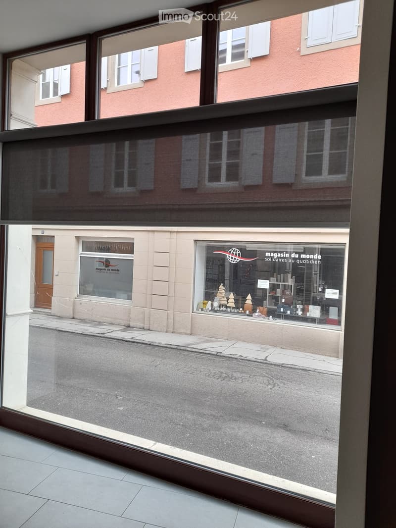 Balcony overlooking a street with a shop named "magasin du monde" visible in the reflection. The balcony has large glass panels and a wooden frame.