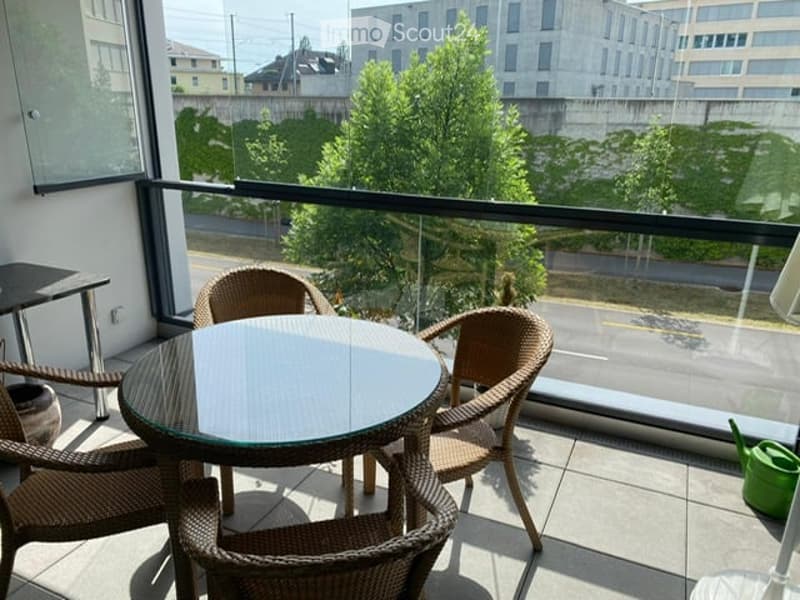 Balcony with a round glass table and four wicker chairs, overlooking a cityscape with trees and buildings.