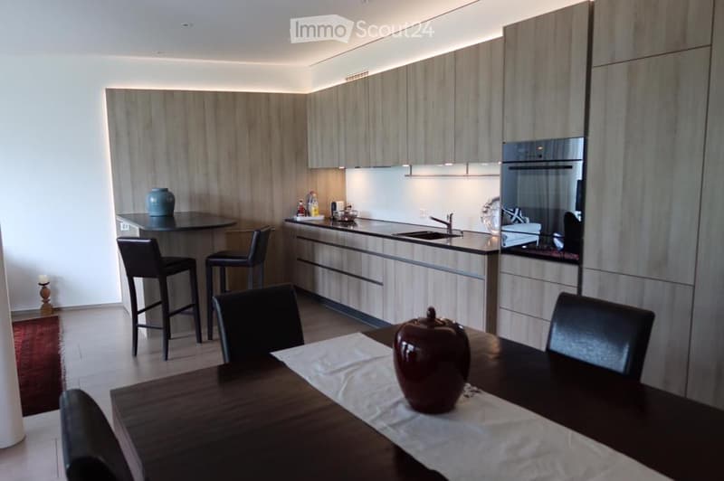Modern kitchen and dining area with sleek wooden cabinets, bar table with stools, dining table with chairs, and a decorative fruit centerpiece.