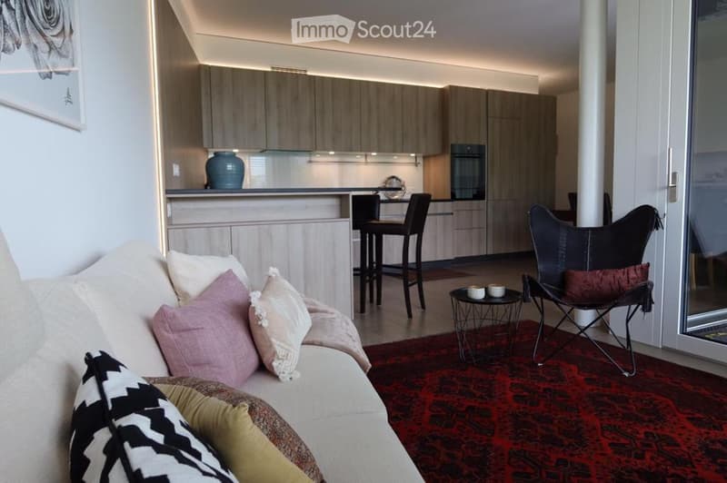 A modern, open-plan living and dining area with a beige sofa, colorful pillows, a black leather chair, a small round table, and a dining area with bar stools and a kitchen counter.