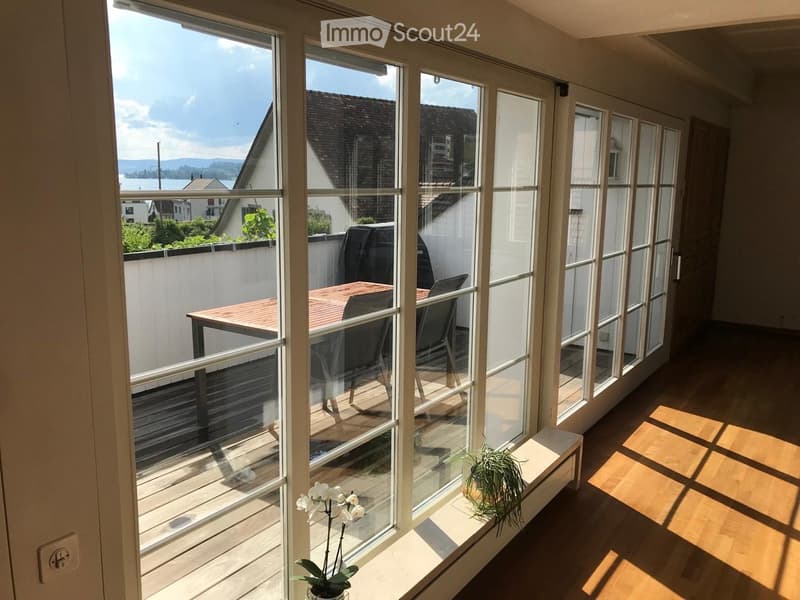 Bright, sunlit balcony with large glass windows, wooden floor, and a small table with chairs.