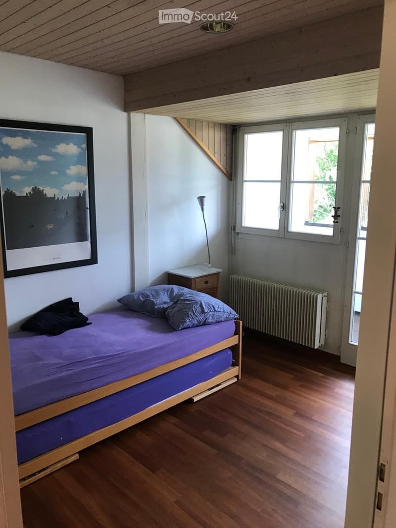 Cozy attic bedroom with wooden slanted ceilings, hardwood floors, a single bed with purple bedding, a nightstand with a lamp, and a radiator near the window.
