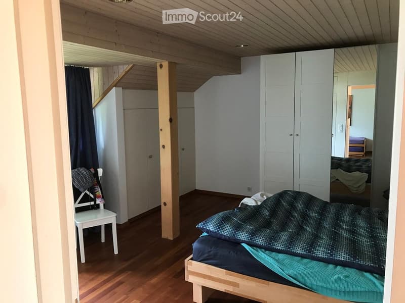 Cozy bedroom with wooden floors, a bed with blue bedding, a white chair, and a large wardrobe. The room features exposed wooden beams on the ceiling and a dark curtain on one wall.