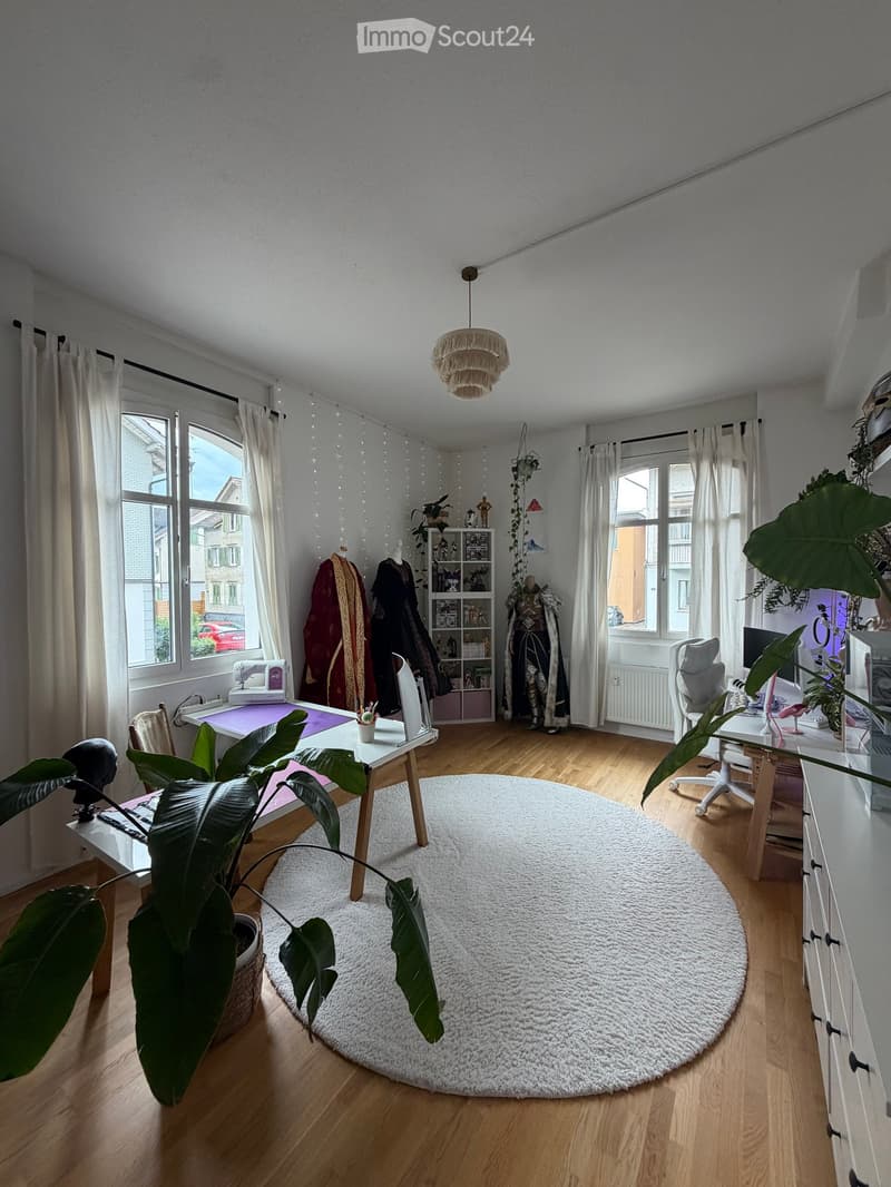 A well-lit room with wooden flooring, large windows, and a round white rug. Contains a table with a plant, a chair, shelves with books and decorative items, and a hanging lamp.