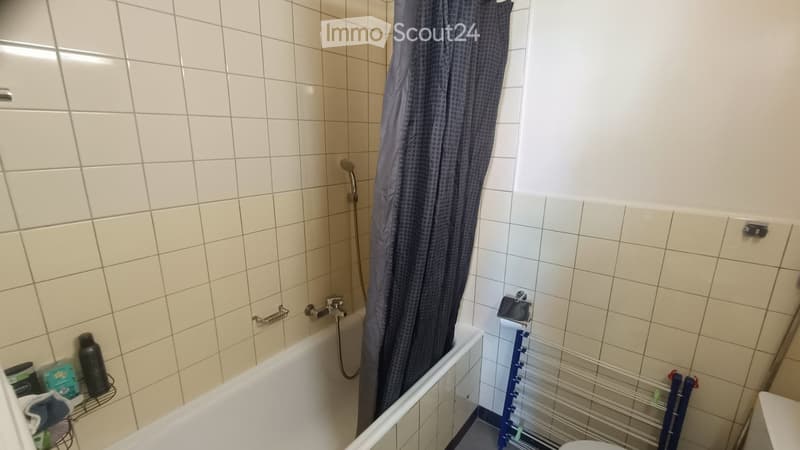 Bathroom with a bathtub, shower curtain, and a towel rack. The walls are tiled, and there is a small shelf with a bottle and other items.