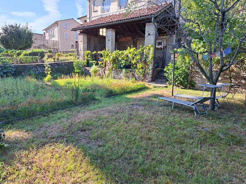 Garden area with a stone wall, grassy lawn, and outdoor furniture including a table and chairs near a tree. A rustic building with a tiled roof is visible in the background.