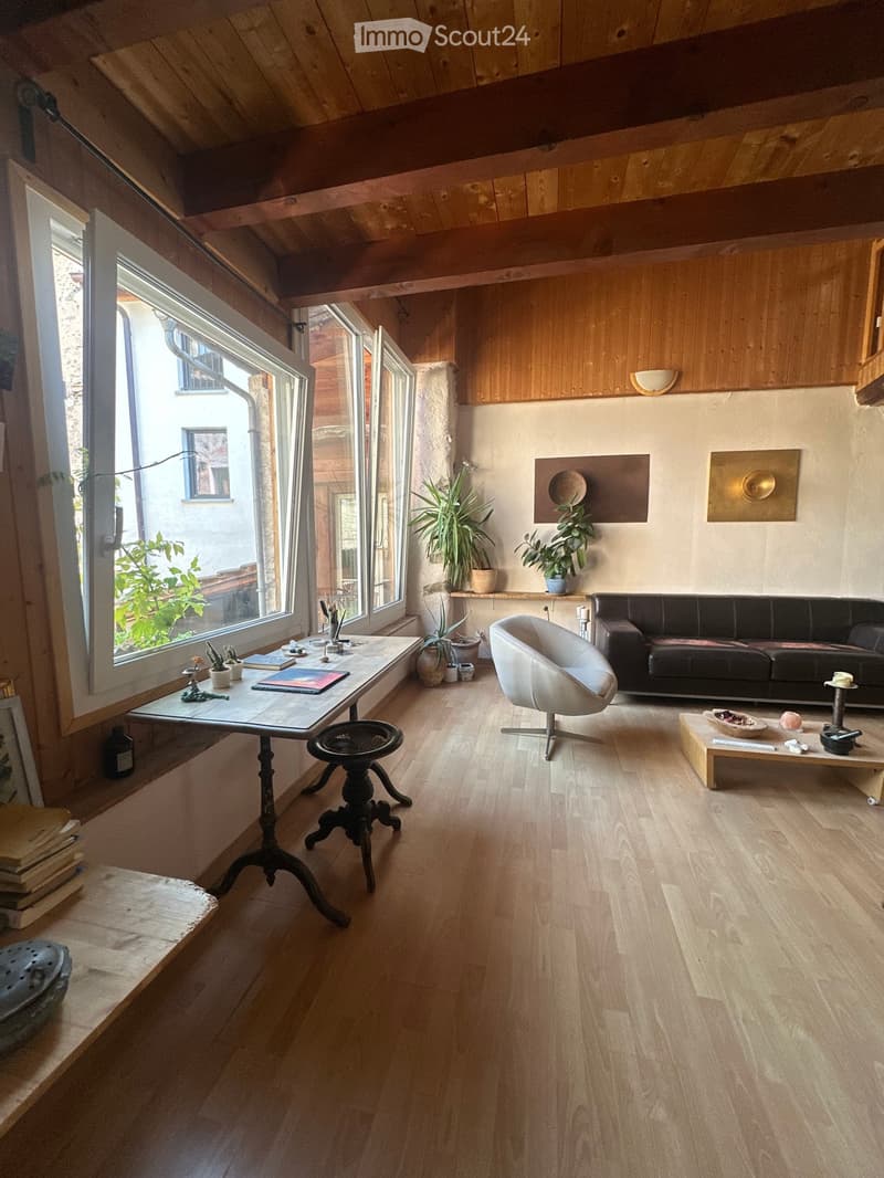 Bright living room with wooden floors, large windows, a black leather sofa, a white armchair, a wooden coffee table, and various decorative items including plants and art pieces.