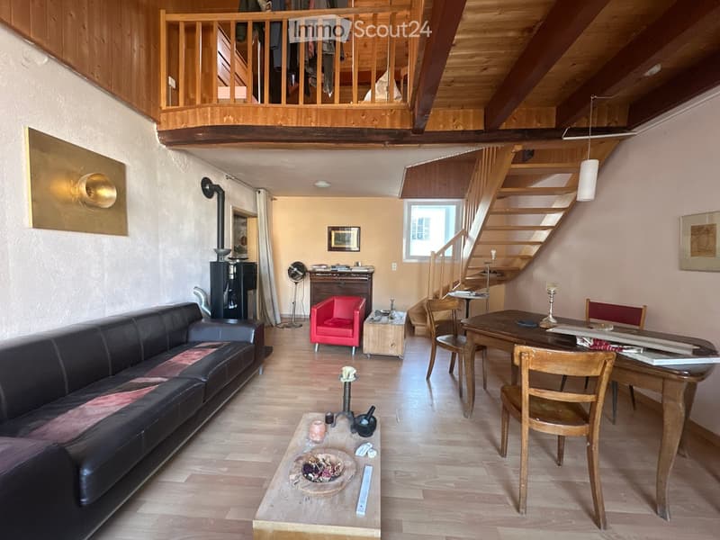 The image shows a cozy, well-furnished living room with wooden floors, a wooden ceiling, and a wooden staircase leading to an upper level. The room features a black leather sofa, a red armchair, a wooden coffee table, a wooden dining table with chairs, a