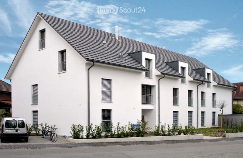 Modern two-story white building with grey roof, multiple windows, balconies, and small garden area in front.