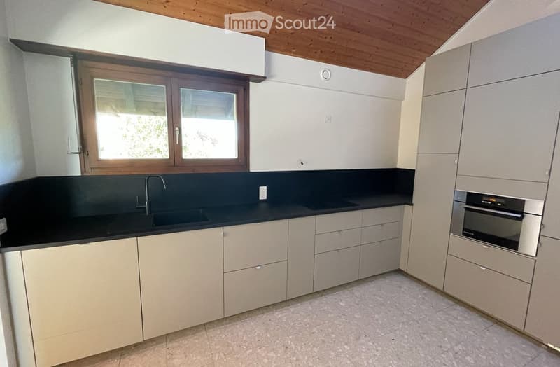 Modern kitchen with light-colored cabinets, black countertops, a sink with a faucet, and an oven. The room features a wooden ceiling and a window with wooden frames.